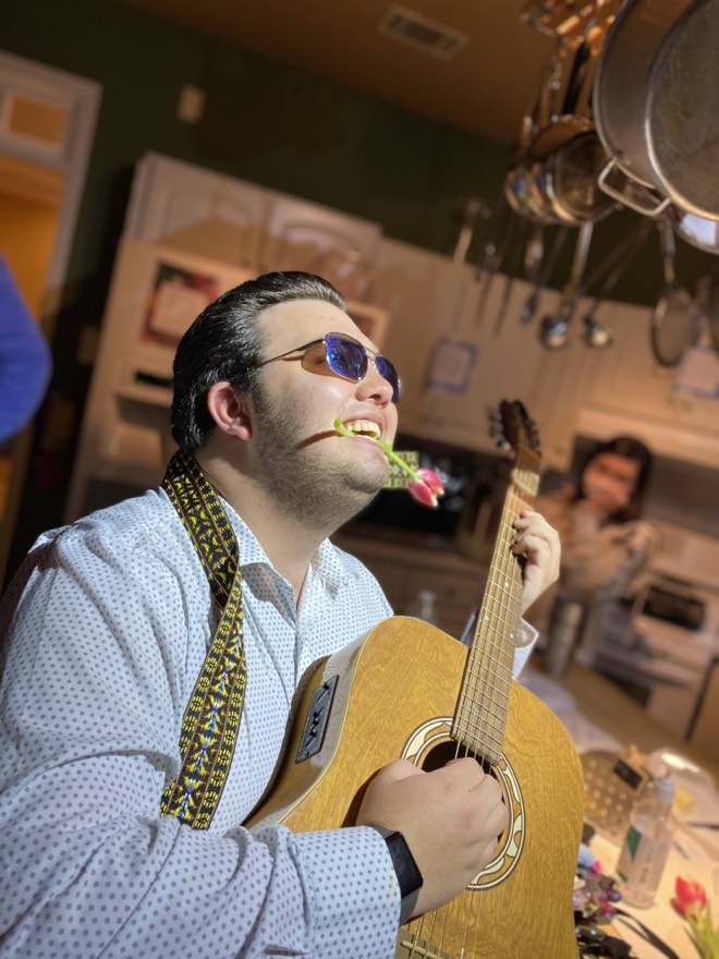 Bibby in sunglasses playing an acoustic guitar with a tulip in his teeth, in a kitchen at night