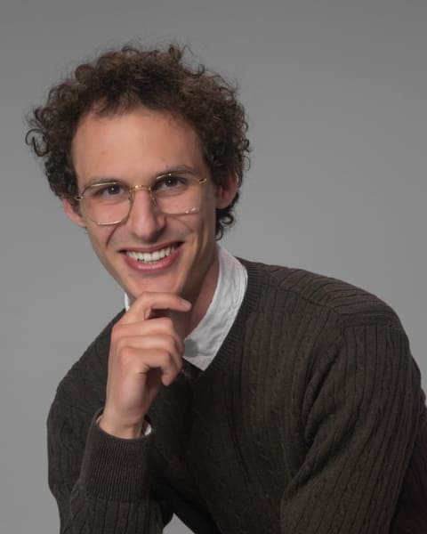 Grinning young man in glasses and a knit sweater, chin resting on his hand.