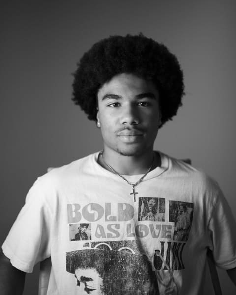Black-and-white portrait of a young man with an afro and a small cross necklace.