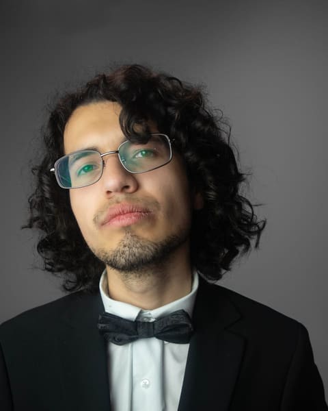 Close portrait of a man with long curly hair, wire-rimmed glasses and a bow tie.